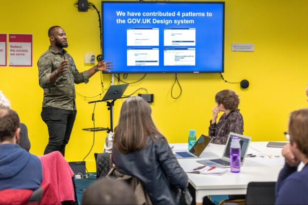 A man giving a talk in front of several people. There is a screen on the wall behind him that reads 'We have contributed 4 patterns to the GOV.UK Design System'.