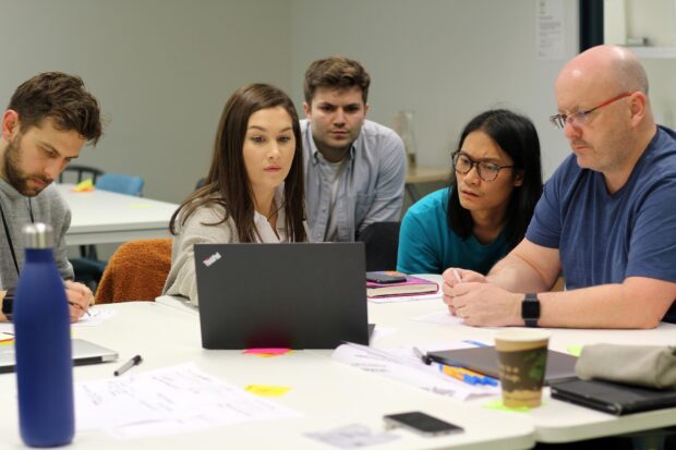 5 people sitting around a table and looking at a laptop, one of them is taking notes