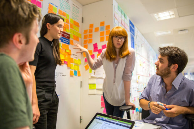 Two young female and two male designers discuss ideas in front of a wall with colour stickie notes; one holds a laptop computer, another one has a sketchpad in front of him