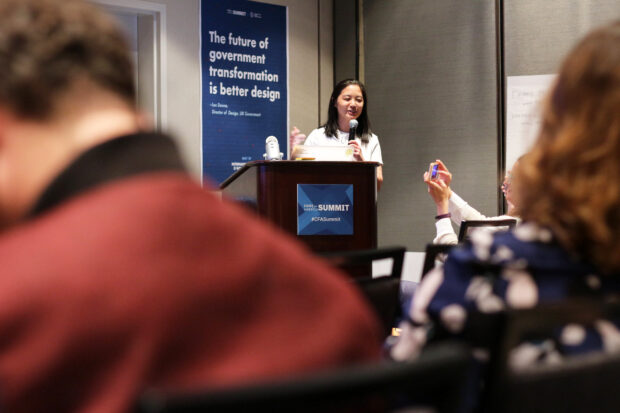 Mari Nakano, Design Director at the NYC Mayor's Office for Economic Opportunity, presenting in front of a group of people at the International Design in Government Day in Oakland; a poster behind her reads: “The future of government is better design”; the speaker smiles and members of the audience take pictures with their smartphones