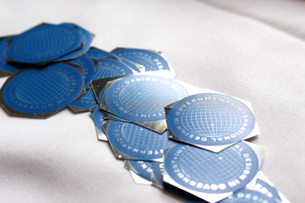 A dozen blue stickers with an illustration of the globe and the words ‘International Design in Government’ on a white table cloth