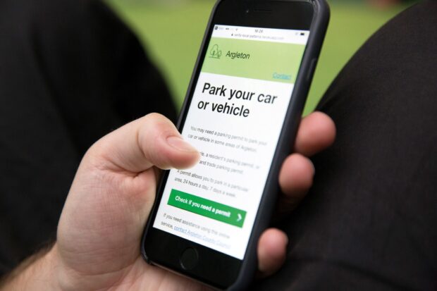 A close-up photo of a man’s hand holding a smartphone; it shows a local government service for parking your car or vehicle of the fictional local council of Argleton