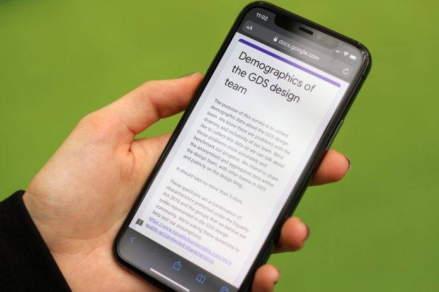 A hand of a person with light skin holding a smartphone showing an online survey; its headline says: ‘Demographics of the GDS design team’