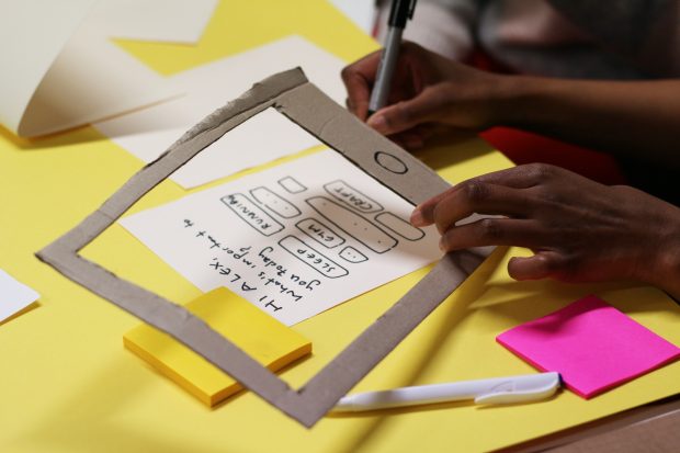 A woman with darker skin tone is preparing a mockup of a smartphone interface, the phone is a cardboard frame, the screen is small sheet of paper, the person is drawing the user interface with a pen, coloured sticky notes are surrounding her