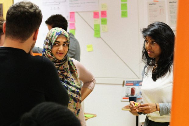 A small of group of people working together during the Gov Jam, in focus: two young women of different ethnicities, one holding a LEGO figure, coloured sticky notes are behind them on the wall