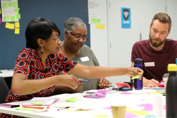 Three participants in GDS service design training – two women with a darker skin, one man with light skin tone working together at a table with coloured sticky notes