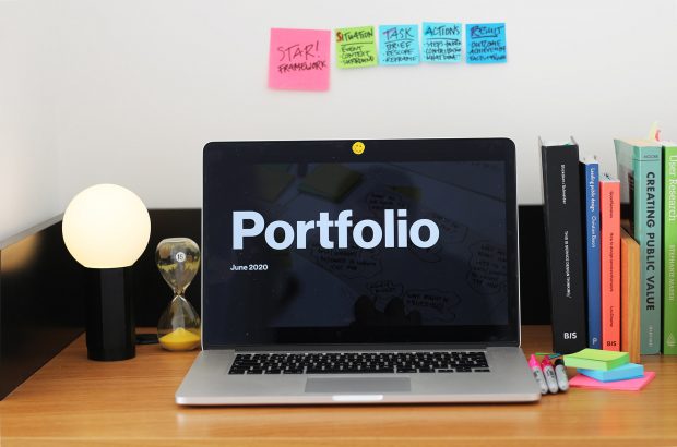 A laptop computer on a wooden desk, its screen shows the title page of a design portfolio