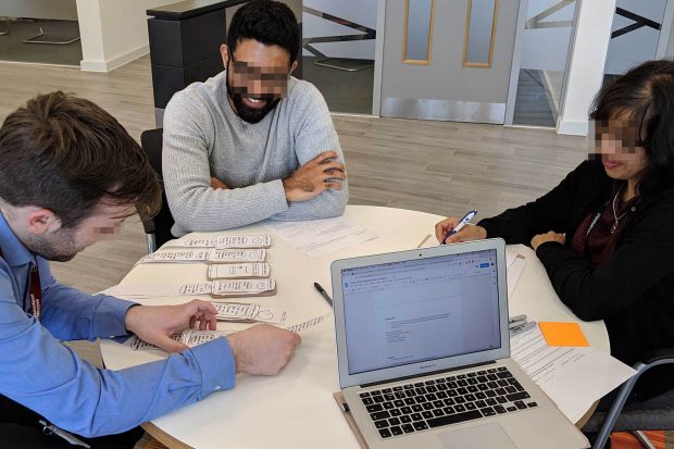 Three people of different ethnicities smiling, sitting around a table in a co-creation workshop looking at paper prototypes of mobile service interfaces