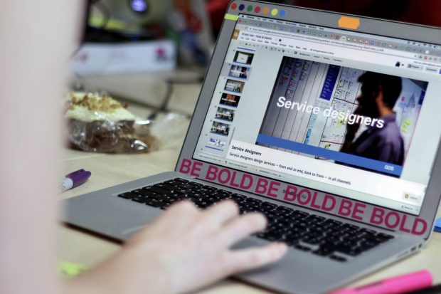 A person working on a laptop computer that shows one slide of a presentation: the slide says ‘service designers’ and shows a person in the background who looks at a wall full of notes, the slide is branded as GDS, and the laptop has various BE BOLD stickers on it