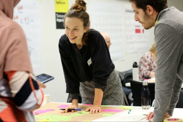 A group of people standing over a service blueprint draft, the central person, a young woman, holding a pen and rearranging stickie notes