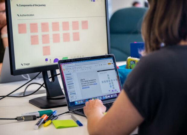 Image of a person sitting on a desk, next to pens and post-its notes, in front of an opened laptop screen and a monitor behind it