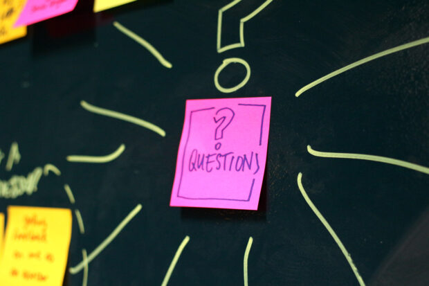 A post it on a wall, it says 'questions? on it, and participants might be about to stick post its beneath it