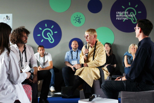 A group of 4 people with microphones and lanyards are sitting on stools, discussing. 4 more people who look at them sit behind them. There is a wall with graphical applications of logos of ‘Creative Bureaucracy Festival’. All people have lighter skin tones.