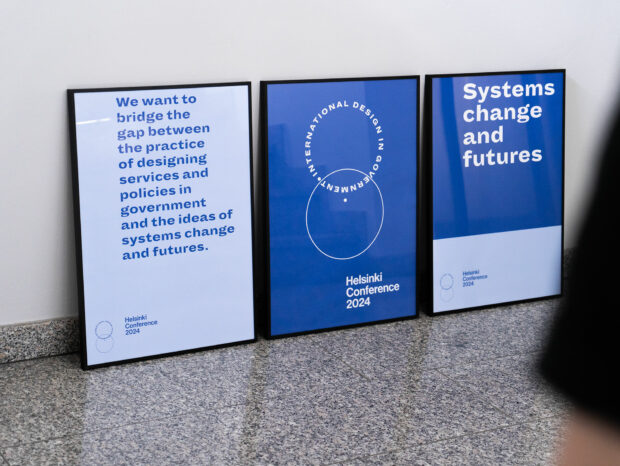 Three framed posters for the Helsinki Conference 2024 in a corridor – one showing the logo of the International Design in Government community, one saying ‘Systems change and futures’ and one containing a mission statement: “We want to bridge the gap between the practice of designing services and policies in government and the ideas of systems change and futures.”