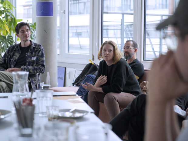 A group of four people sits around a table in a casual meeting. A woman in the center is speaking, while the others listen attentively. The setting is a bright room with large windows and indoor plants.