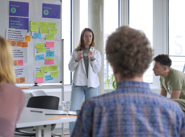 A woman stands in front of a whiteboard filled with colourful sticky notes, presenting to a group. Two individuals listen attentively. The whiteboard has two sections labeled "Group 2" and "Group 3" with various questions and comments written on the notes. The room is bright with large windows in the background.