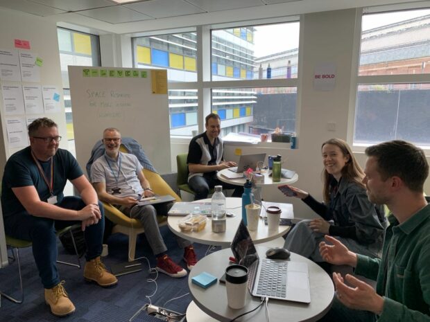 image of the heads of design working group sitting in a group around small tables with our laptops smiling at the camera in a government office in Manchester.