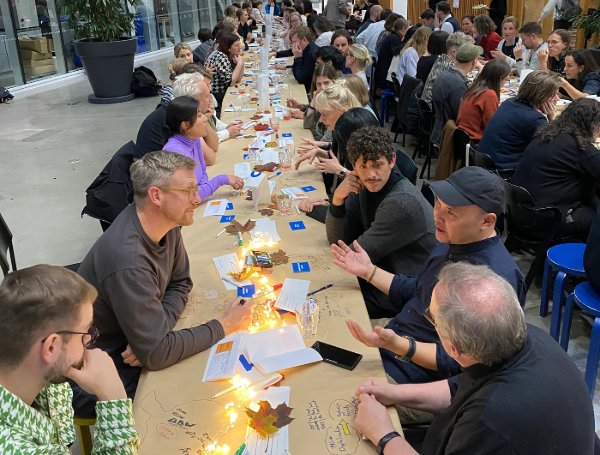 ALT: “People are seated at a long table in an indoor setting, engaged in discussions. The table is decorated with fairy lights, paper notes, and autumnal leaves. The group appears collaborative and focused.”
