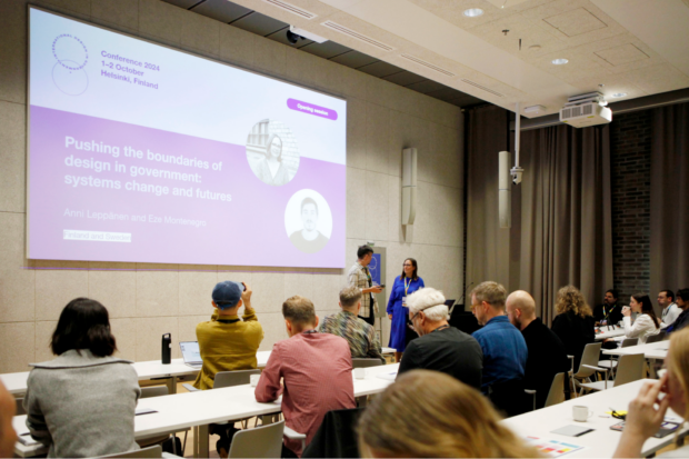 “A conference room with attendees seated at tables, watching two speakers on stage. A large screen displays the presentation title, ‘Pushing the boundaries of design in government: systems change and futures,’ and photos of the presenters. The room has modern decor with a projector and a curtain-covered wall.”