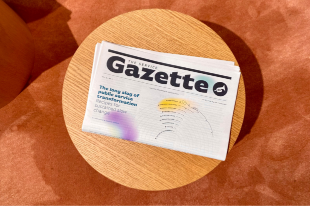 A once-folded newspaper – ‘The Service Gazette’ – on a small circular wooden table; the cover story is: “The long slog of public service transformation: Recipes for sustained slow change”.