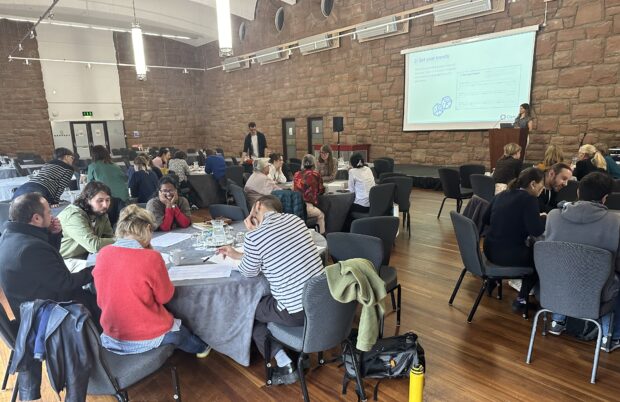 A big room at a conference with a speaker, Benny at a podium in front of a screen presenting. Filling the room are 6 round tables with 5 people sitting around each. Each table is looking at A3 papers and speaking within their groups.