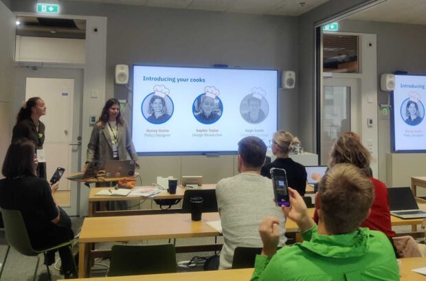 Sophie and Benny standing by a screen presenting their work at the Helsinki Conference. The screen is showing their intro slide with images of themselves and their names. In the room are people sitting at long desks listening to the presentation. 