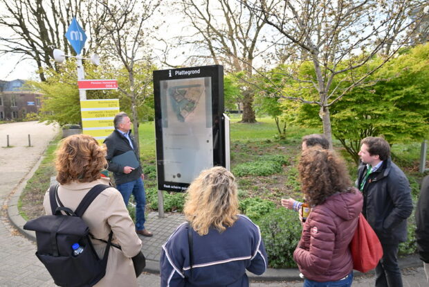 A group of 6 people – men and women – standing outdoors, looking at a large map display mounted on a stand. They appear to be looking at the map and discussing the content.  Trees and a building are in the background.