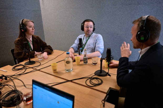 Three people, one woman and two men, sitting around a wooden table in a small, soundproofed space, all wearing big headphones and have professional microphones in front of them.