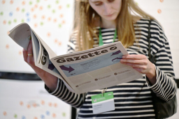 A woman wearing a striped black and white jumper and a conference lanyard, holding two copies of a Service Gazette newspaper and reading the content.